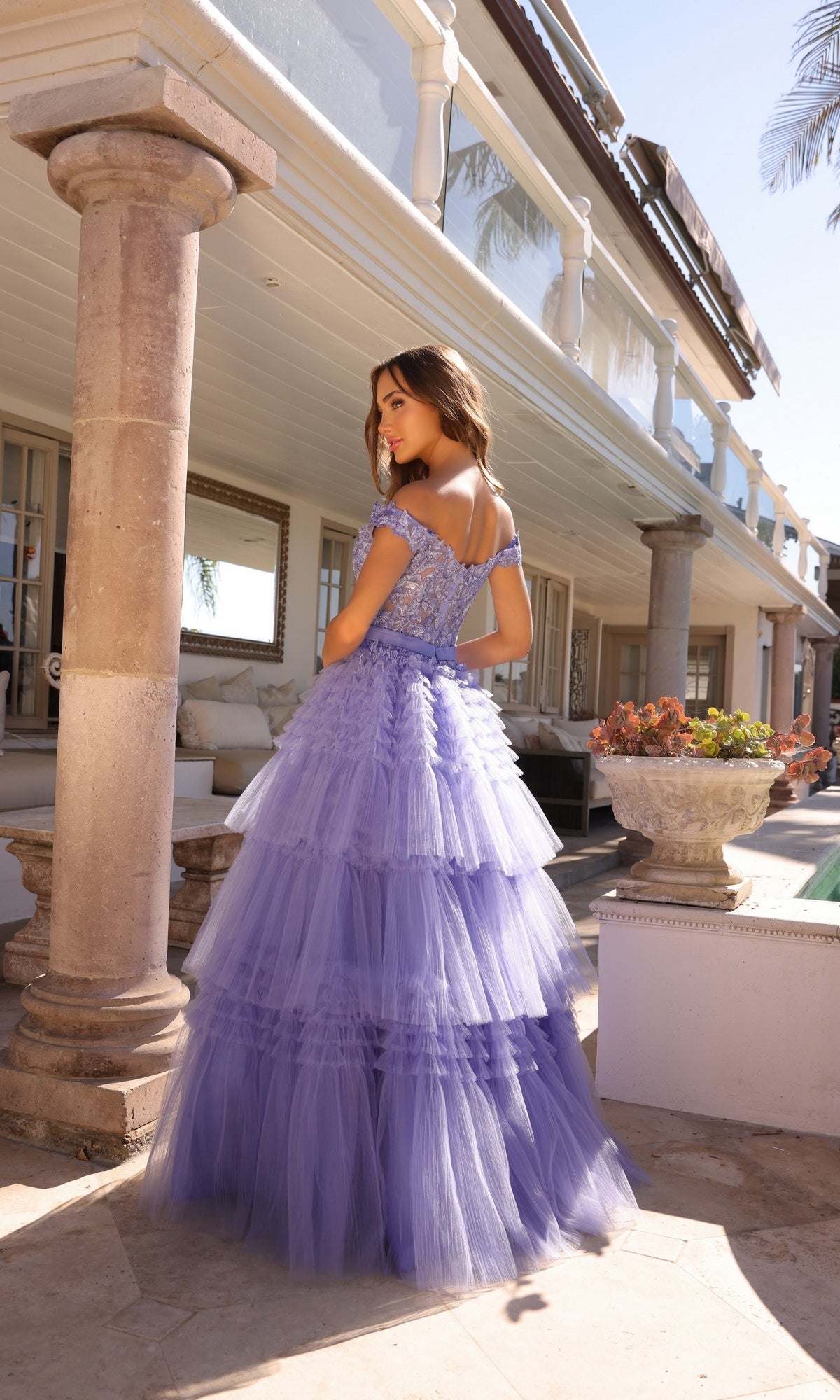 A woman stands outside a building, looking over her shoulder in the Nox Anabel Designer Ruffled Prom Ball Gown E1293—an off-the-shoulder, layered lavender tulle gown with beaded embroidered lace details.